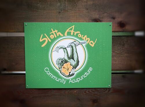 A green sign reads Sloth Around Community Acupuncture.  In the center of the sign is a drawing of a three toed sloth hanging from an acupuncture needle.  The sloth holds an orange hibiscus flower in its right hand.  The sign has rain drops on it, and is nailed to three horizontal wooden boards.  